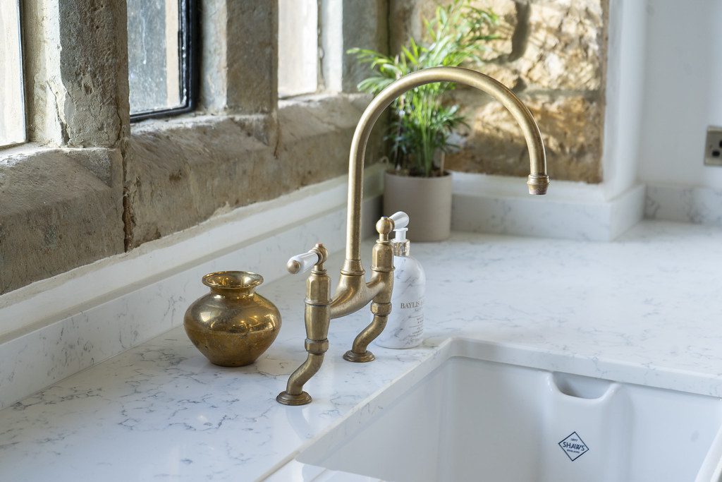 A close-up of a white quartz countertop with subtle grey veining, featuring a vintage-style brass kitchen tap, white ceramic sink, and stone-framed window in a rustic kitchen.